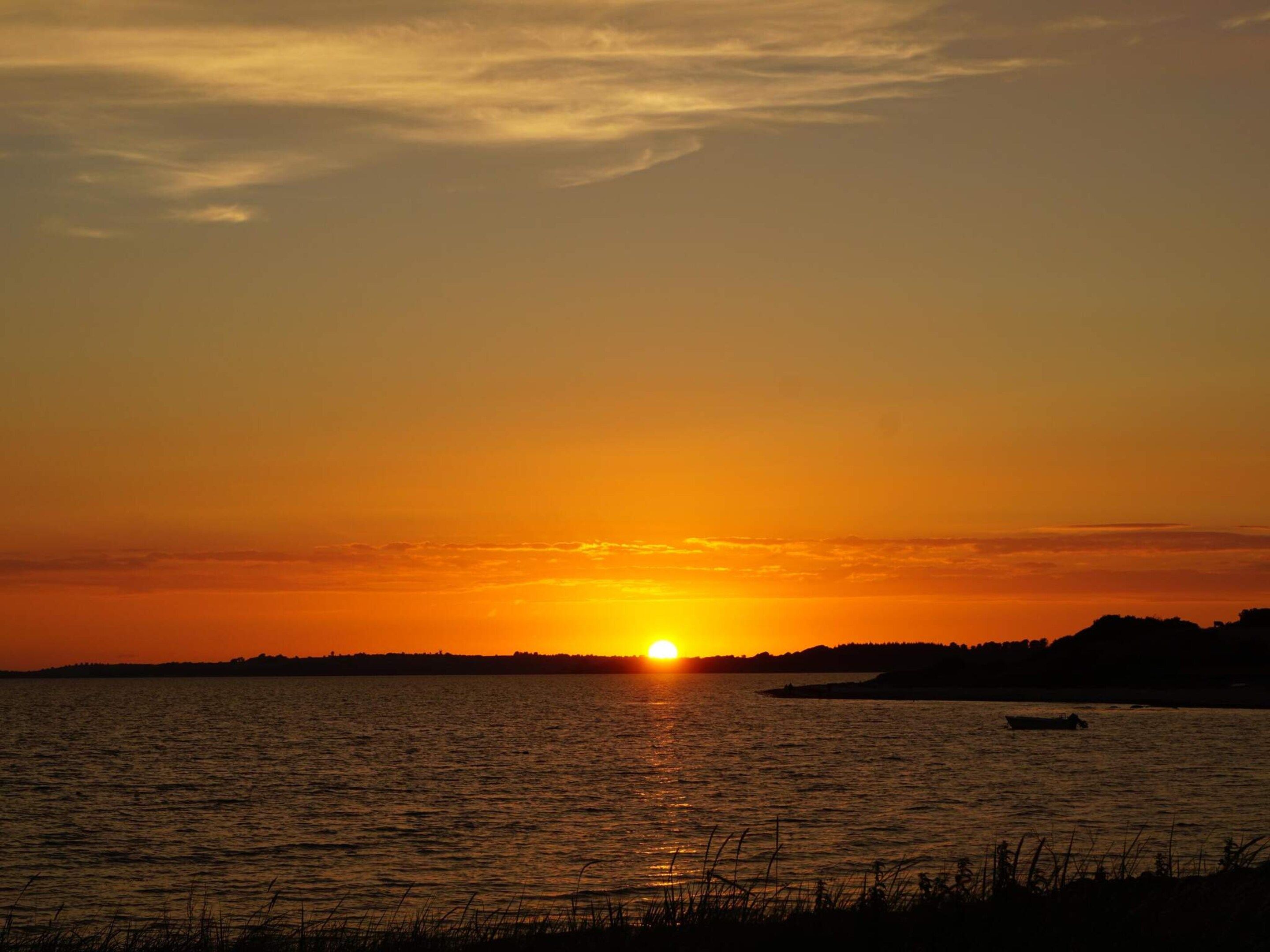 Ein Sonnenuntergang über einem Gewässer, mit der Silhouette eines Bootes und weiter Landschaft im Hintergrund.