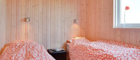 A room with two beds, wooden walls, a window with a flower, and two wall-mounted lights.