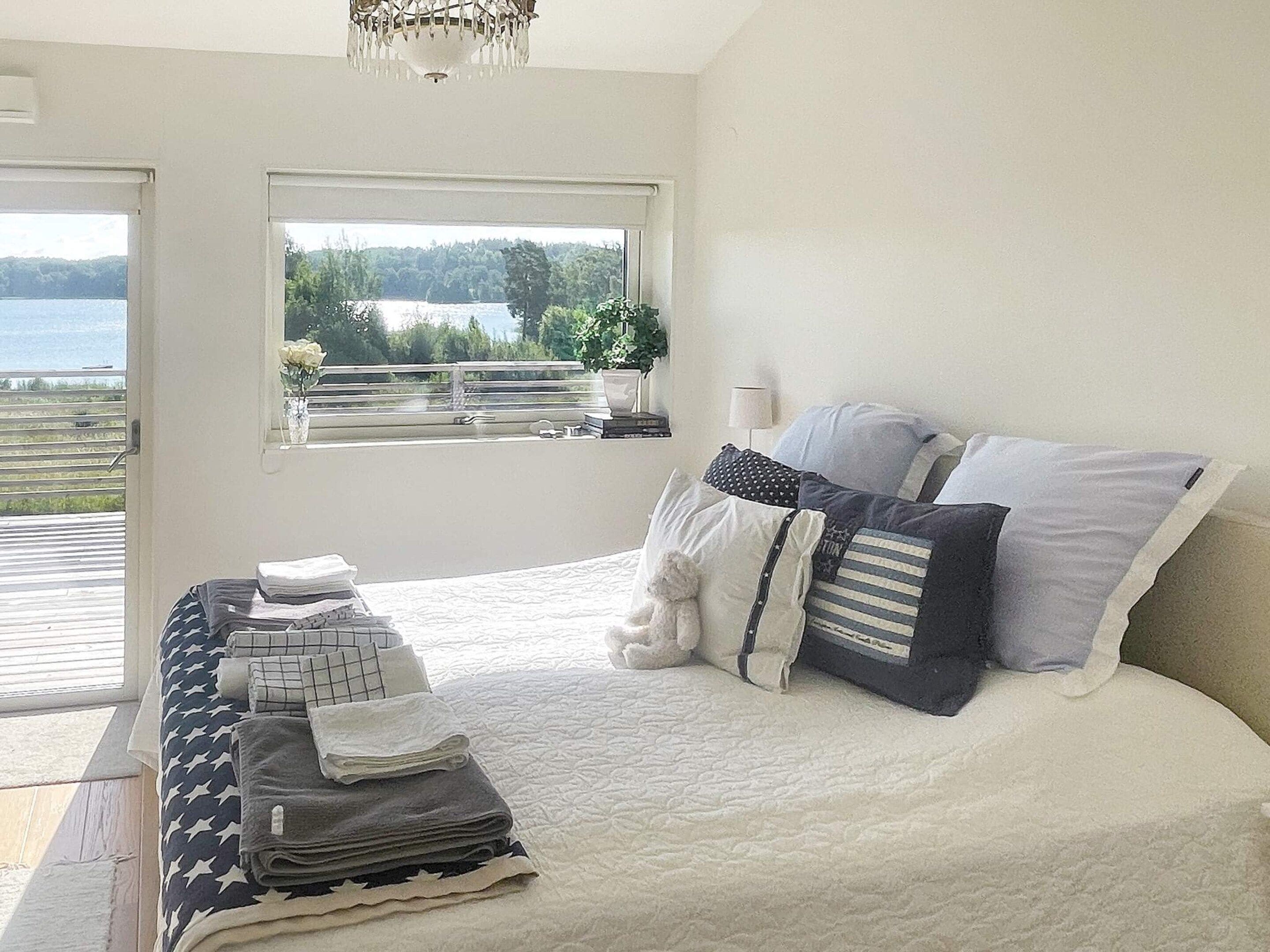 A bedroom with a bed, pillows, a bedside table with folded towels, a window with a view of a lake and trees, and a chandelier.