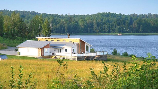 A lakeside cabin with a deck, surrounded by a forested area and a clear sky. - Holiday Home in Stjärnhov (Stjarnhov)