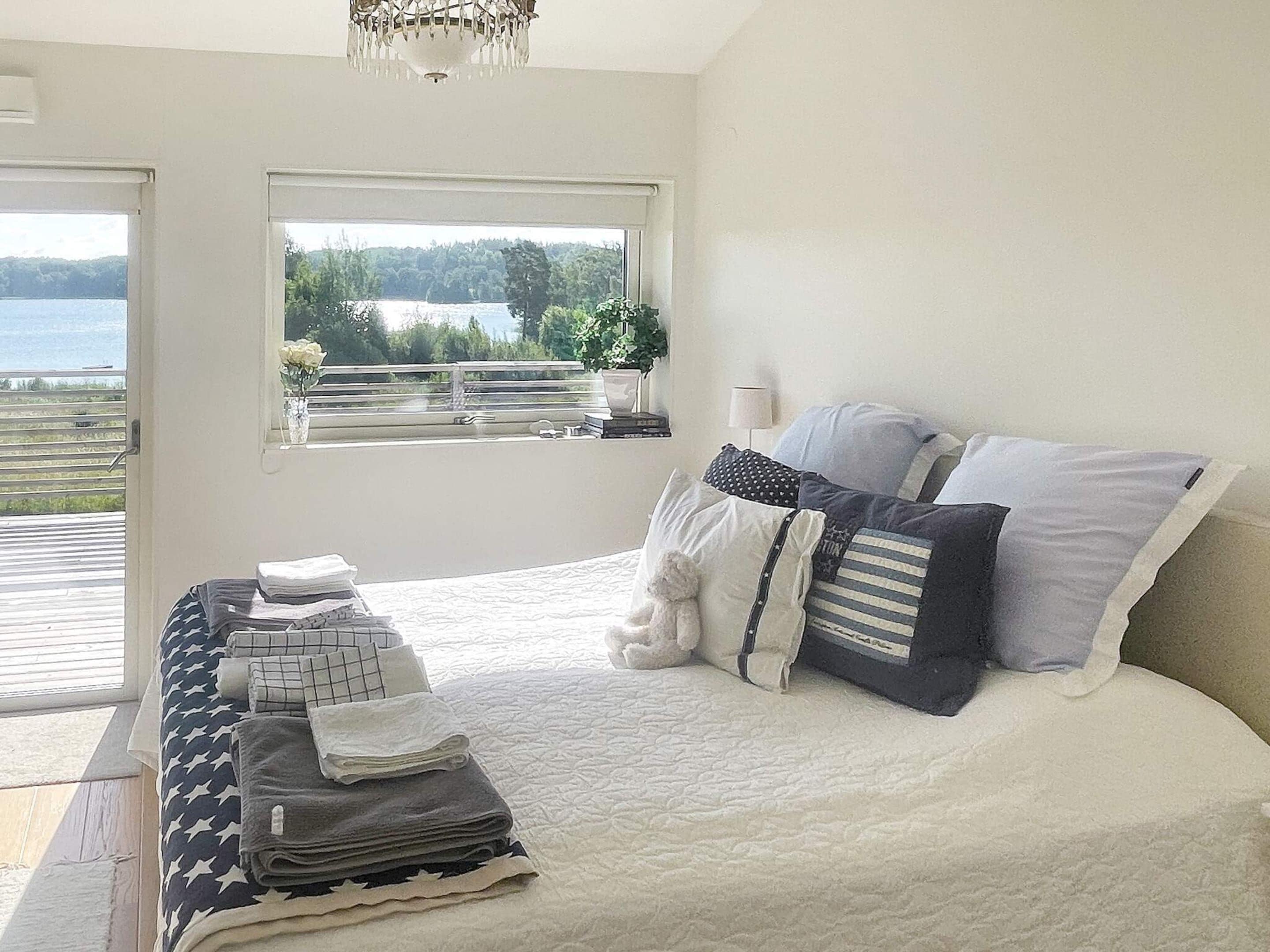A bedroom with a bed, pillows, a bedside table with folded towels, a window with a view of a lake, and a chandelier.