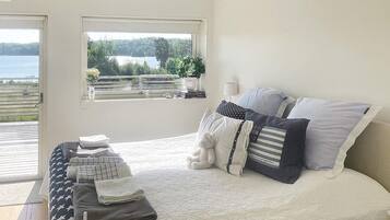 A bedroom with a bed, pillows, a bedside table with folded towels, a window with a view of a lake, and a chandelier.