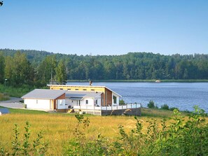 Uma cabana à beira do lago, com varanda, rodeada por uma floresta e um céu claro.