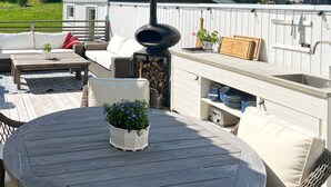 A round wooden table with a potted plant, surrounded by wicker furniture and a built-in grill station. - Holiday Home in Stjärnhov (Stjarnhov)