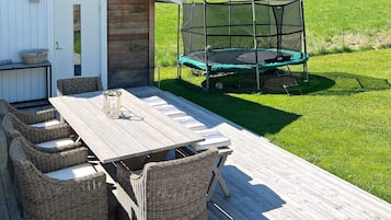 An outdoor deck with a wooden table and wicker chairs, a trampoline, and a covered play area.