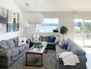 A modern living room with grey sofas, a wooden coffee table, and a large window offering a view of a lake. - Holiday Home in Stjärnhov (Stjarnhov)