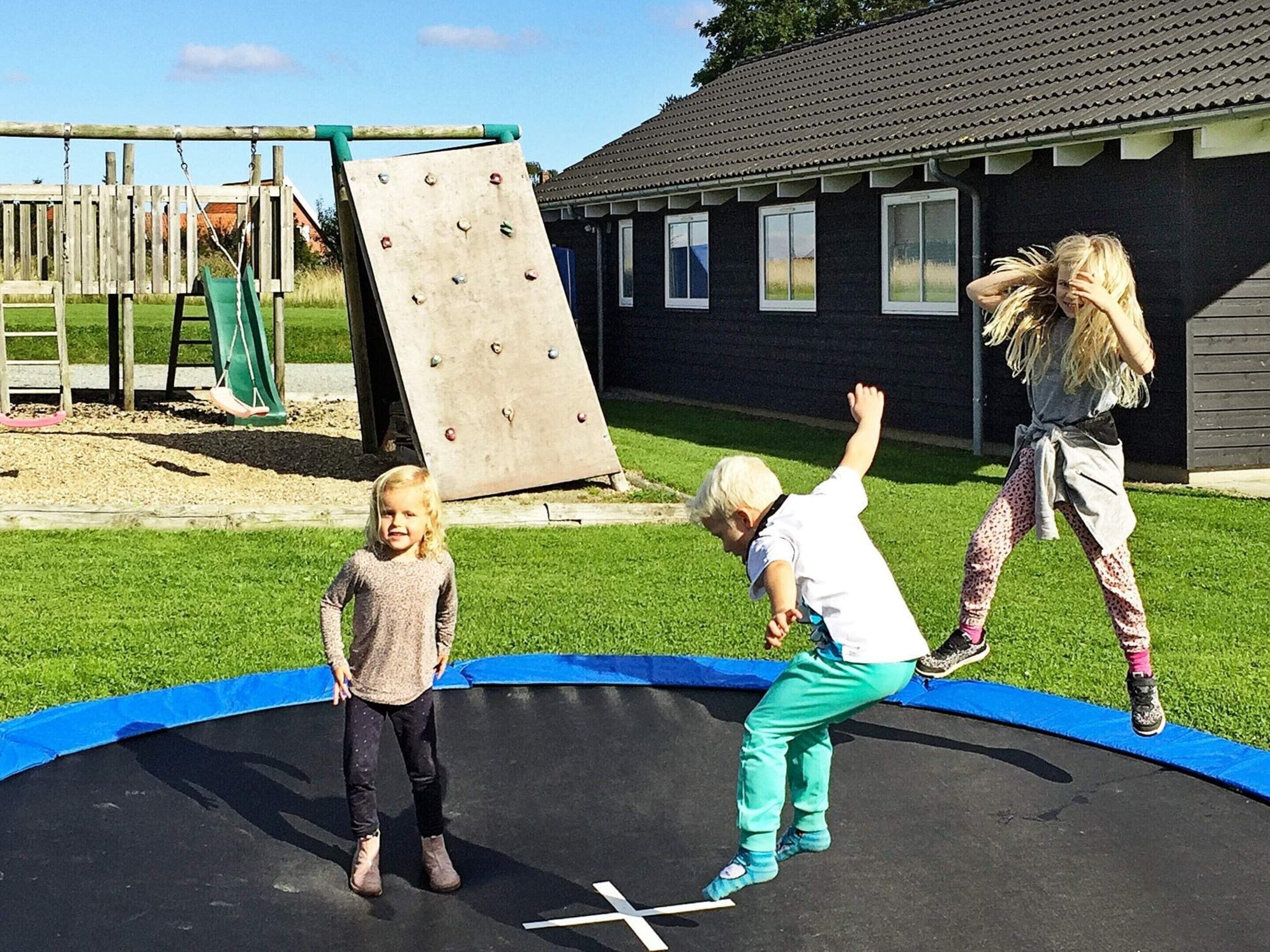 A trampoline with children playing, surrounded by a wooden play structure and a building with windows.