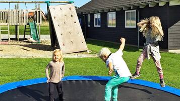A trampoline with children playing, surrounded by a wooden play structure and a building with windows.