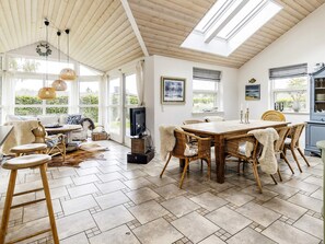 A spacious room with a high ceiling, a dining area with a wooden table and wicker chairs, a television, and a view of greenery outside. - 4 Star Holiday Home in Vaeggerlose (Vaeggerlose)