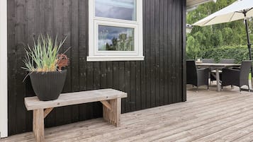 Una terraza de madera con un banco, una planta en maceta, una ventana y mobiliario exterior.