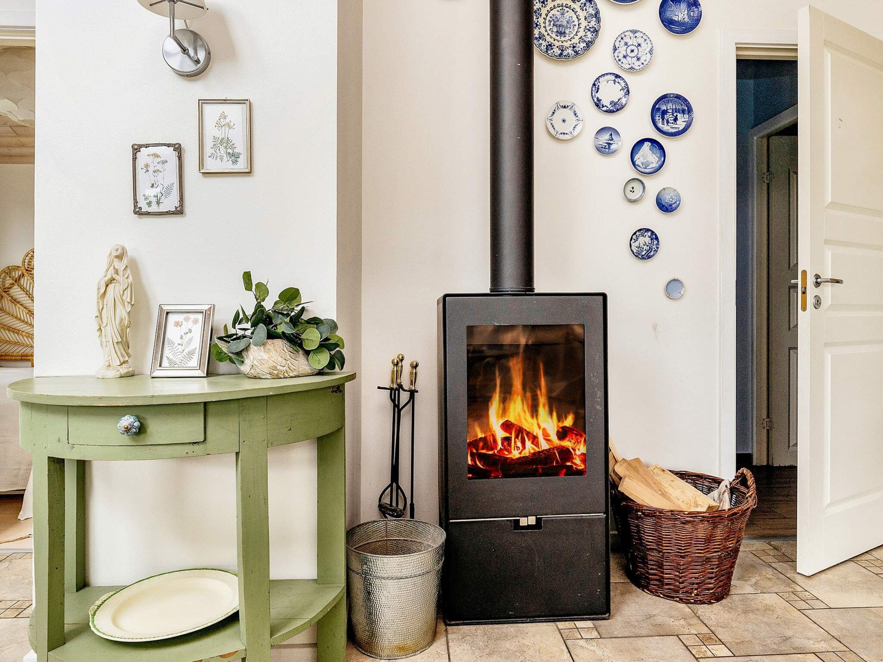 A room with a black wood-burning stove, a green table with a plate, and a basket of firewood.