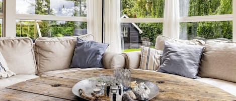 A cozy seating area with a wooden table, cushions, and a view of greenery outside.
