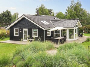 A black house with a tiled roof, a covered patio with outdoor furniture, and a garden with tall grass. - 4 Star Holiday Home in Vaeggerlose (Vaeggerlose)