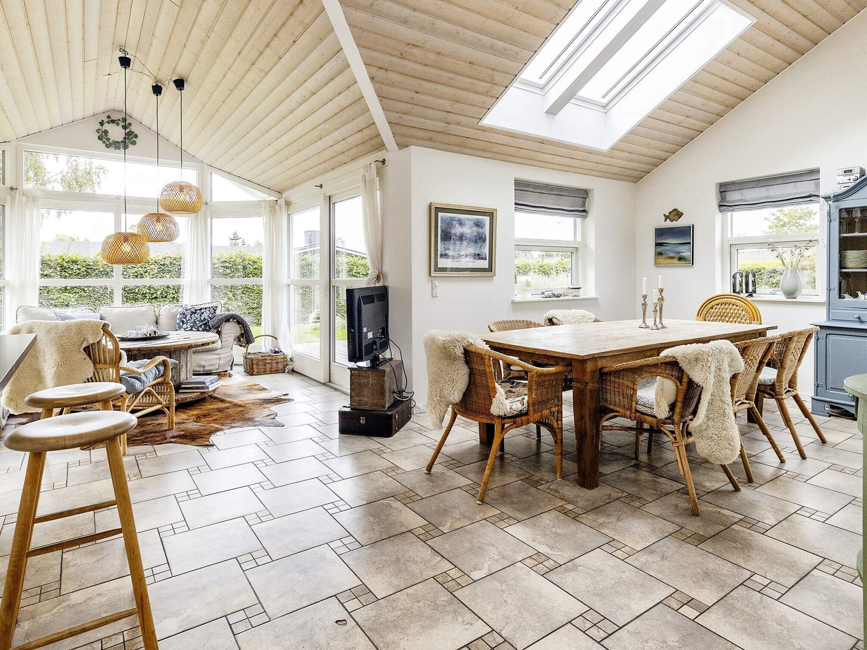 A spacious room with a high ceiling, a dining area with a wooden table and wicker chairs, and large windows allowing natural light.