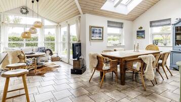 A spacious room with a high ceiling, a dining area with a wooden table and wicker chairs, and large windows allowing natural light.