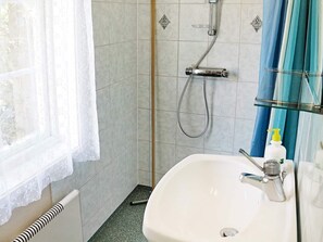 Une salle de bain avec un lavabo blanc, une douche équipée d’un pommeau flexible et une fenêtre ornée d’un rideau léger.