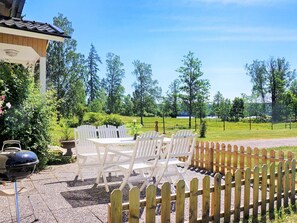 An outdoor seating area with white chairs and a table, surrounded by a wooden fence, with a grill and a pergola in the background. - Holiday Home in Hova (Hova)
