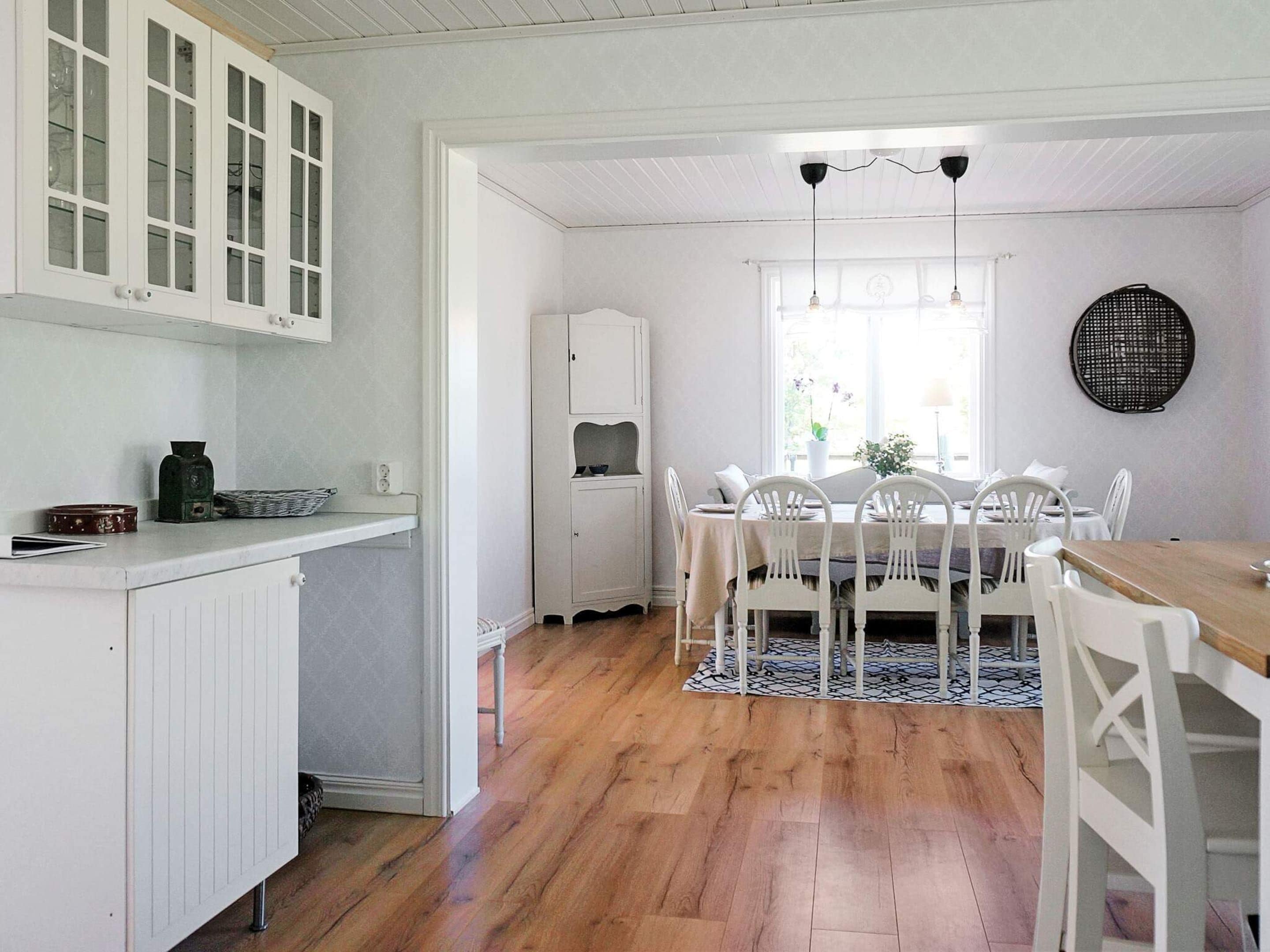 A dining area with a wooden table, white chairs, and a patterned rug. There is a kitchen area with white cabinets and a countertop.