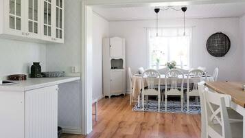 A dining area with a wooden table, white chairs, and a patterned rug. There is a kitchen area with white cabinets and a countertop.