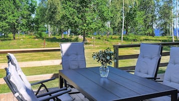 An outdoor dining area with a table and chairs, surrounded by a wooden deck and a view of trees and a body of water.