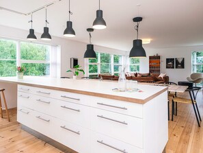 A modern kitchen with a central island, large windows, and a cozy living area with a sofa and bookshelves. - Gorgeous Holiday Home in Ebeltoft with Hot Tub (Ebeltoft)