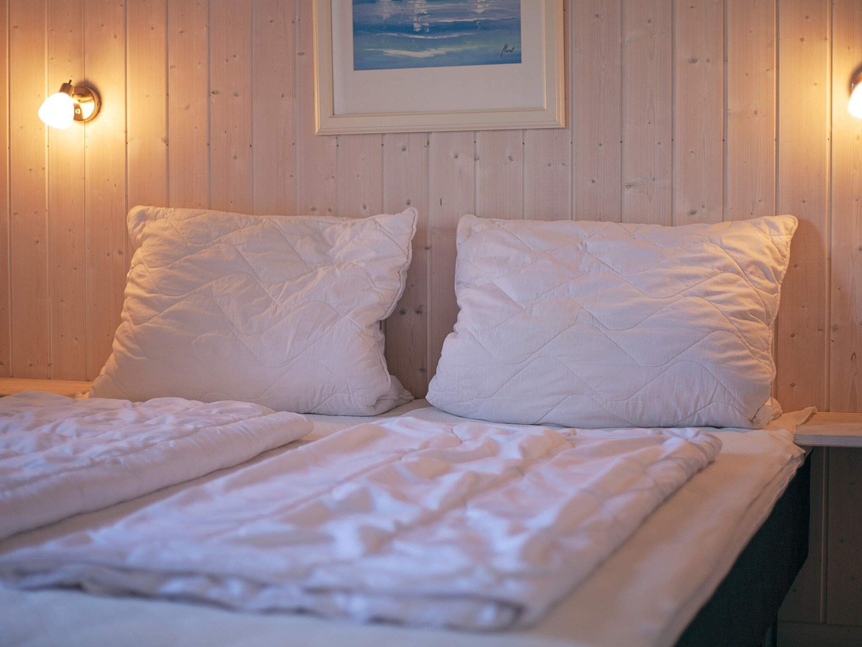A bed with a quilted white duvet and two pillows, a framed picture on the wall, and a wall-mounted light fixture.