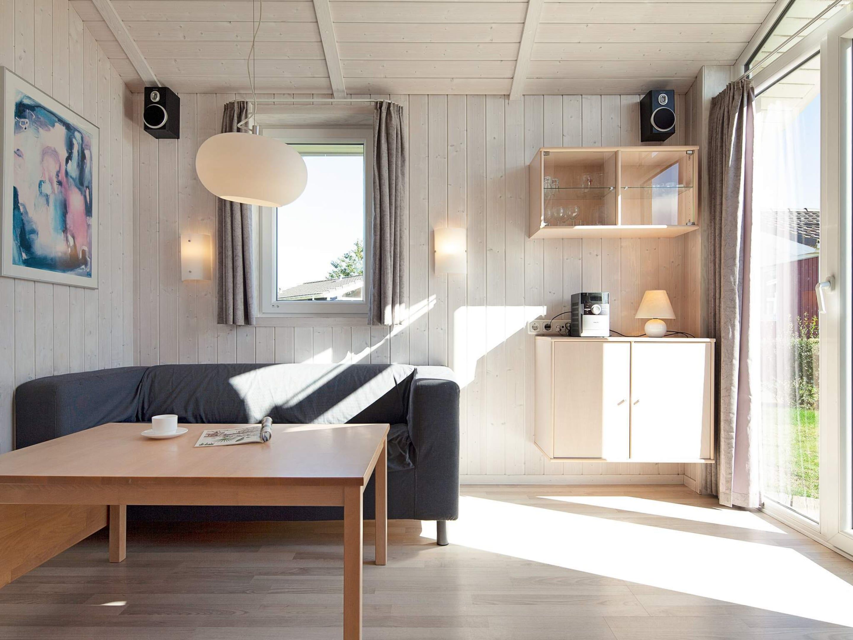 A modern living room with a wooden table, a dark sofa, a pendant light, a kitchen area with cabinets, and a large window with curtains.