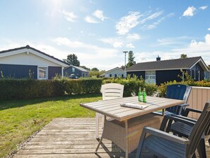 A wooden deck with a table and chairs, surrounded by a well-maintained lawn and a hedge.
