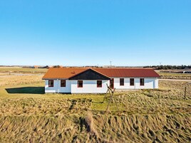 A white building with a red roof and multiple windows, surrounded by open fields.