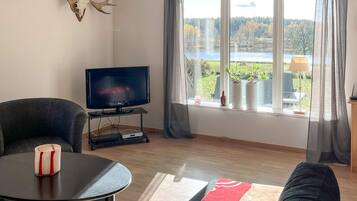 A living room with a television, a sofa, a coffee table, and a window with a view of a lake and trees.