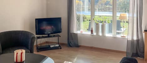 Una sala de estar con televisión, un sofá, una mesa de centro y una ventana con vista a un lago y árboles.