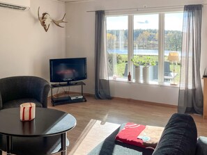 A living room with a television, a sofa, a coffee table, and a window with a view of a lake and trees.