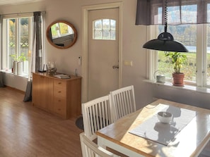 A room with a wooden dining table, white chairs, a wooden cabinet, a round mirror, and a window with a view of greenery.