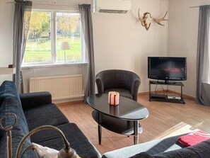 A living room with a sofa, a table with a candle, a TV on a stand, and a window with curtains. - Holiday Home in HÃ¥cksvik (Håcksvik)