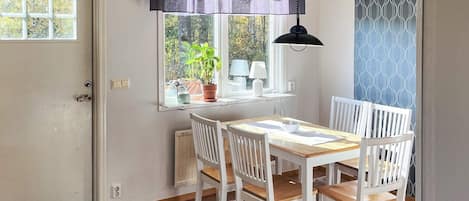 A dining area with a wooden table and white chairs, a window with a view of greenery, and a black hanging lamp.