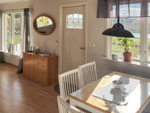 Una habitación con una mesa de comedor de madera, sillas blancas, un armario de madera, un espejo redondo y un ventanal con vista a zonas verdes.