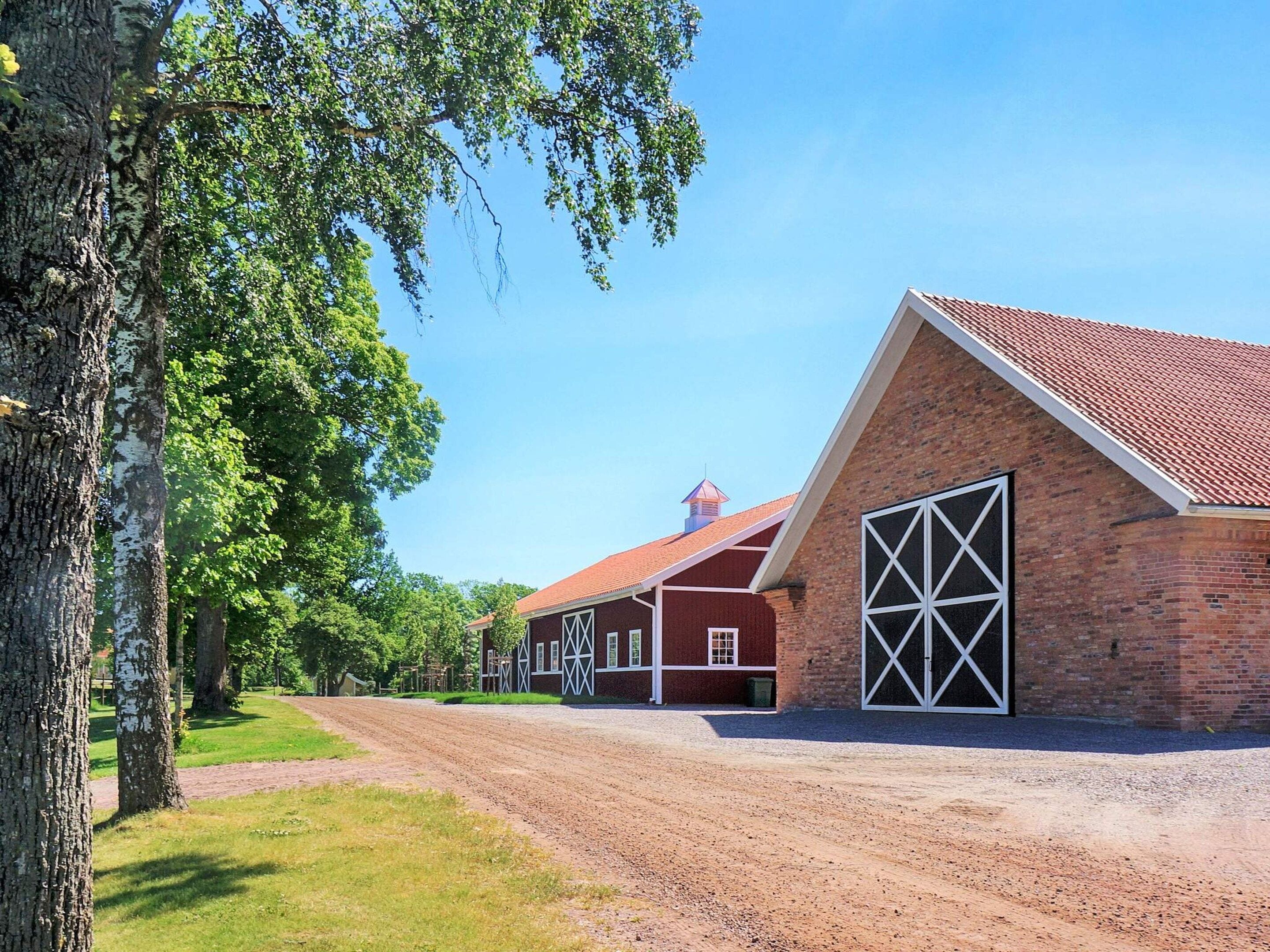 A brick building with a red roof and large white doors, surrounded by trees and a gravel path.