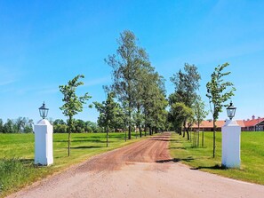Une route de campagne mène à un bâtiment doté d’un toit rouge, bordé d’arbres et de réverbères blancs.