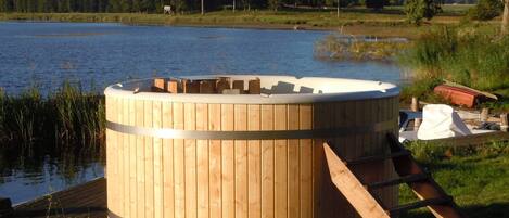 A wooden hot tub with a ladder, situated by a lake with trees in the background.