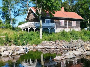 A wooden house with a balcony, surrounded by greenery and a body of water.