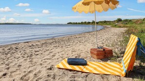 Ein Strand mit einem gelb-gestreiften Liegestuhl, einem passenden Handtuch, einem gepunkteten Sonnenschirm und einem Korbwicker-Korb mit Büchern und Sonnenbrille.