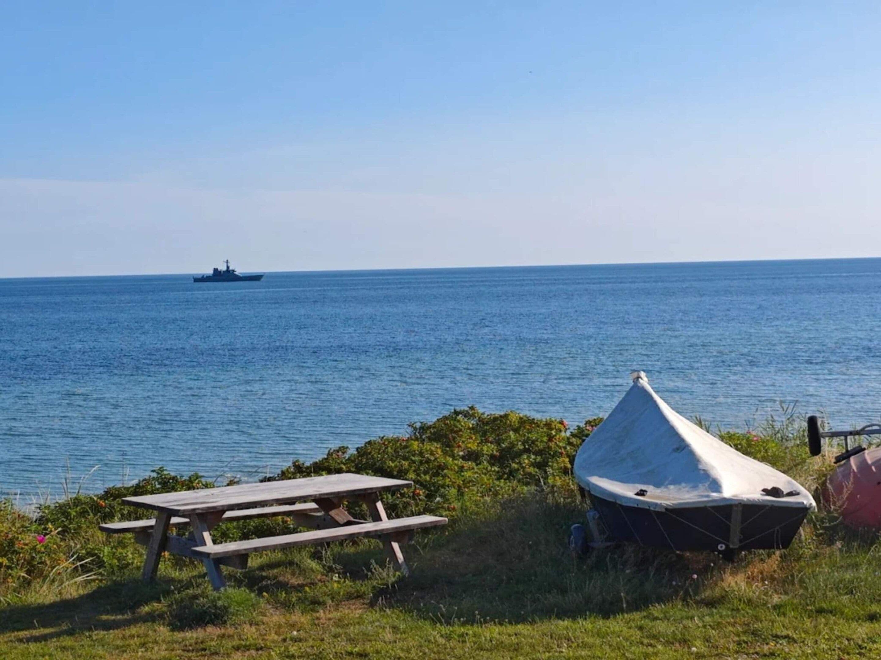 A picnic table and a covered boat in a grassy area with a view of the sea and a ship in the distance.