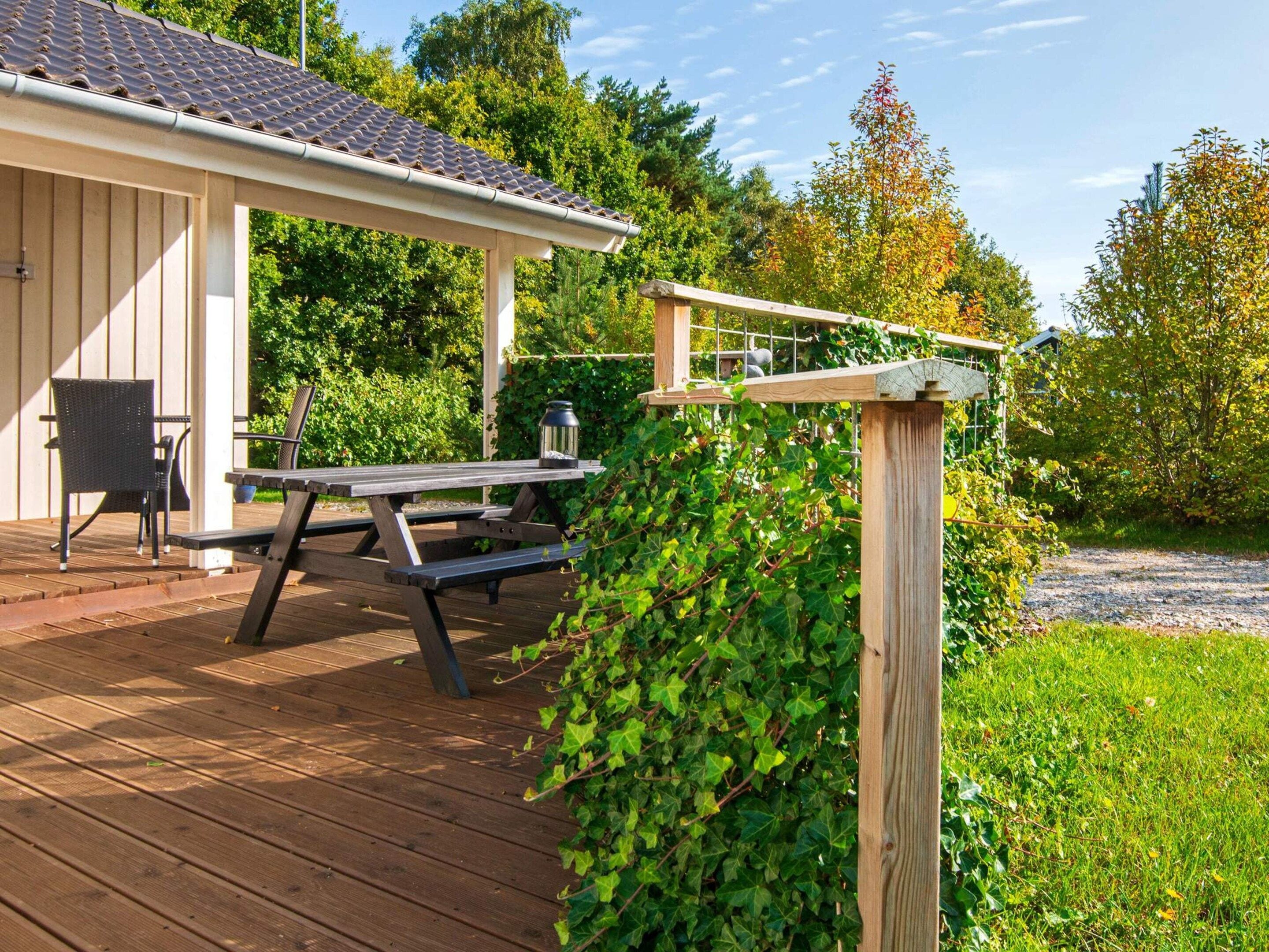Une terrasse en bois avec une table de pique-nique et des chaises, une pergola couverte de plantes grimpantes et, au fond, un bâtiment avec un toit en tuiles.