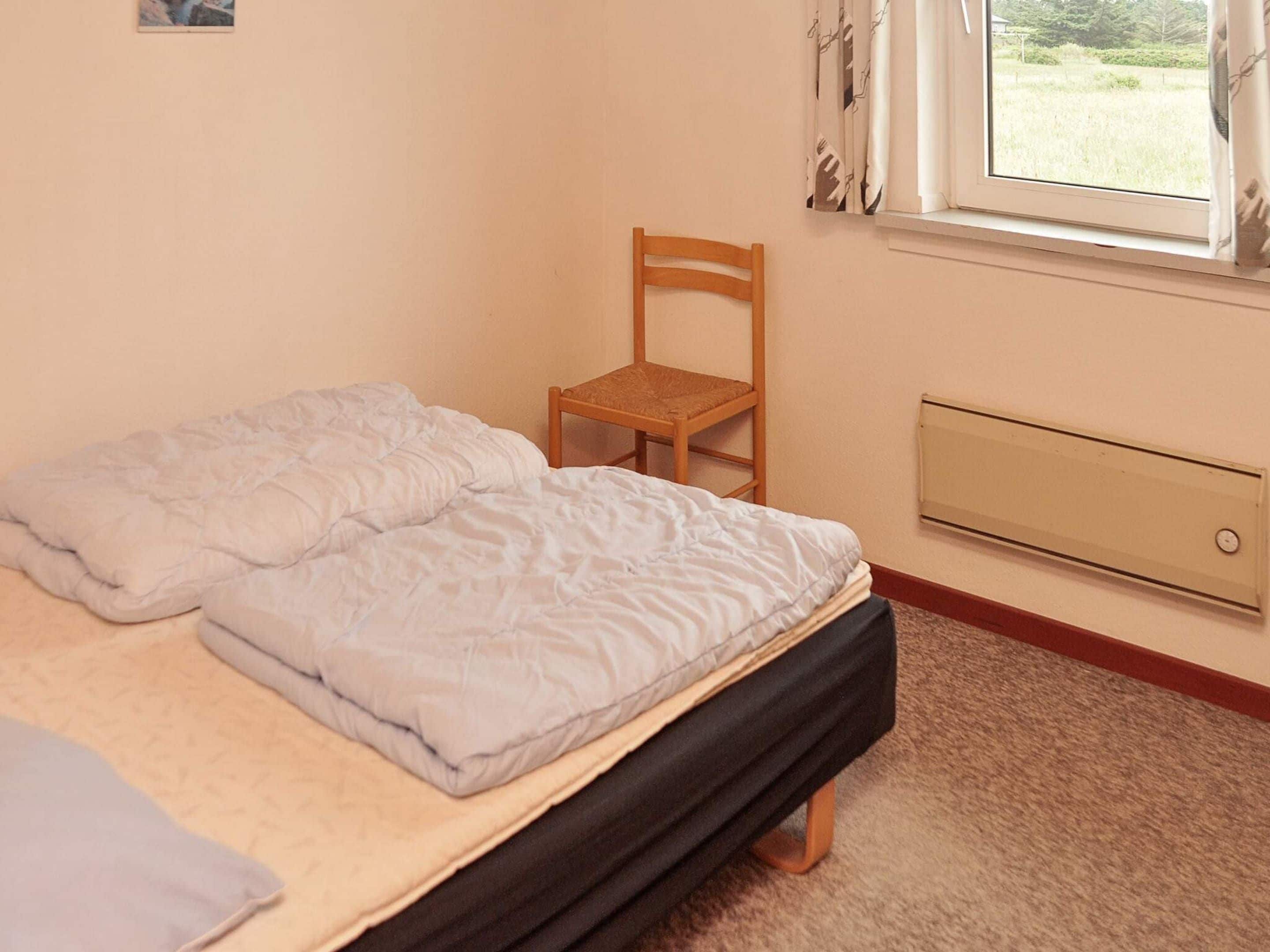 Ein Einzelbett mit Holzrahmen und Matratze, ein Holzstuhl, ein Fenster mit Vorhängen und eine Heizung.