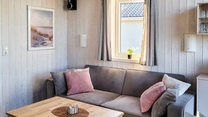 Ein gemütliches Zimmer mit Holzboden, einer Couch mit Kissen, einem kleinen Tisch mit Unterlage, einem gerahmten Bild an der Wand, einem Fenster mit Vorhängen und einem Lautsprecher.