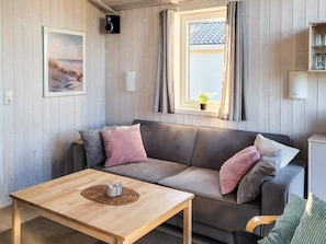 Ein gemütliches Zimmer mit Holzboden, einer Couch mit Kissen, einem kleinen Tisch mit Unterlage, einem gerahmten Bild an der Wand, einem Fenster mit Vorhängen und einem Lautsprecher.