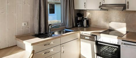 A compact kitchen with a sink, dishwasher, and stove, wooden cabinets, and a window with curtains.