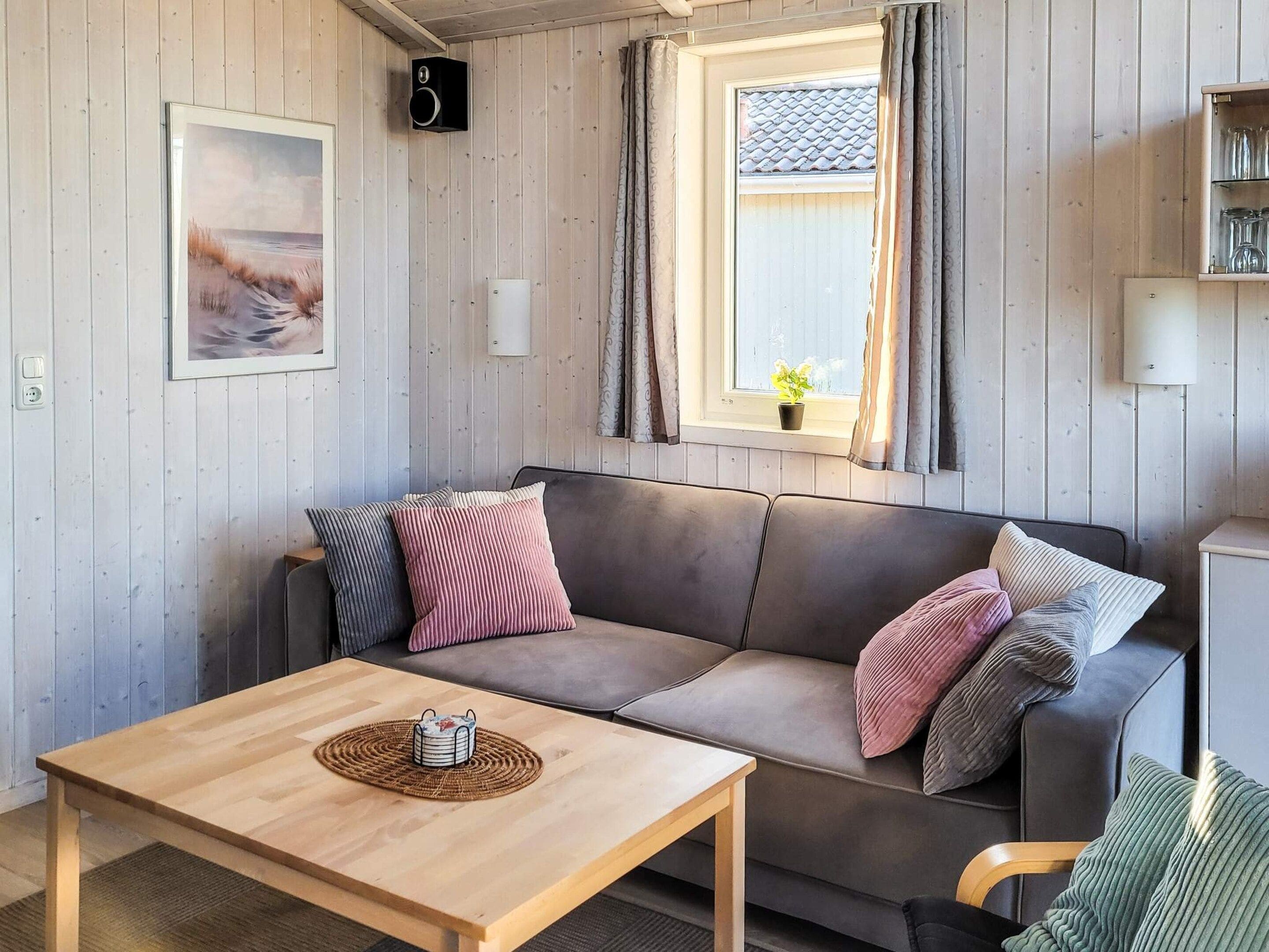 A cozy room with a wooden floor, a grey sofa with cushions, a small wooden table, a framed picture on the wall, a window with curtains, and a speaker.