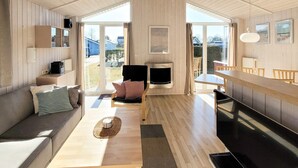 A modern interior with a wooden floor, a sofa, a dining table, a fireplace, and large windows.