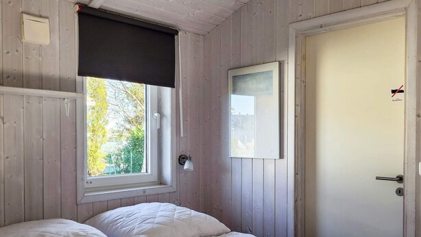 A room with a bed, a window with a dark blind, a door with a sign, and a wall-mounted light fixture.